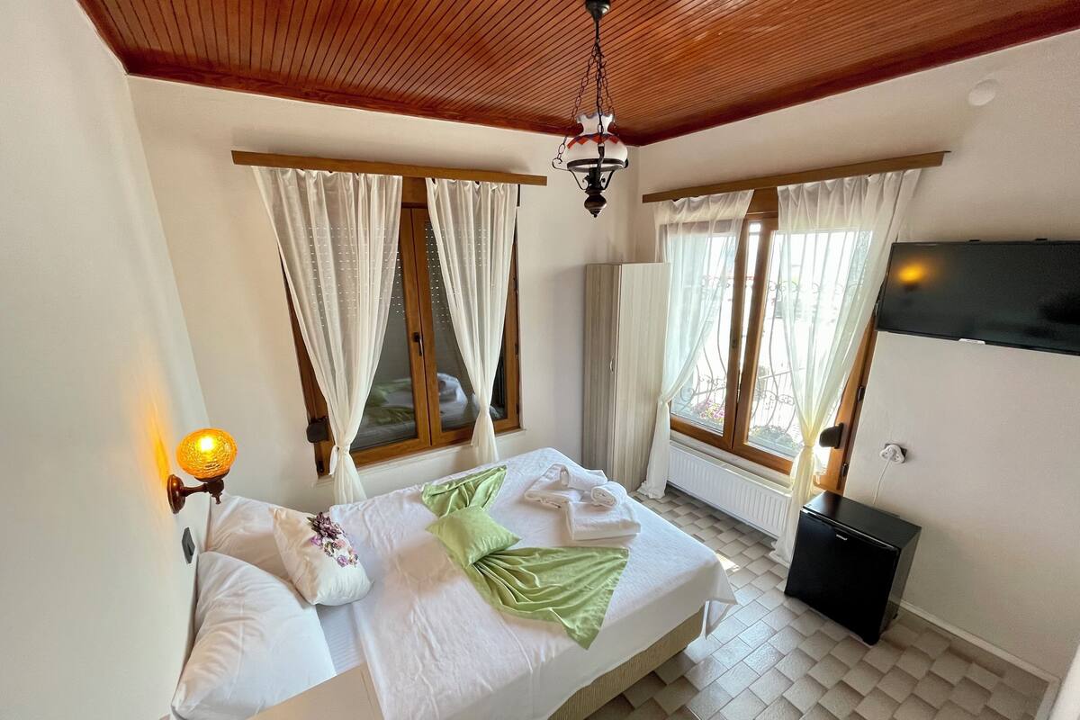 A bedroom with a bed, two windows with curtains, a ceiling fan, a wall-mounted lamp, and a radiator.