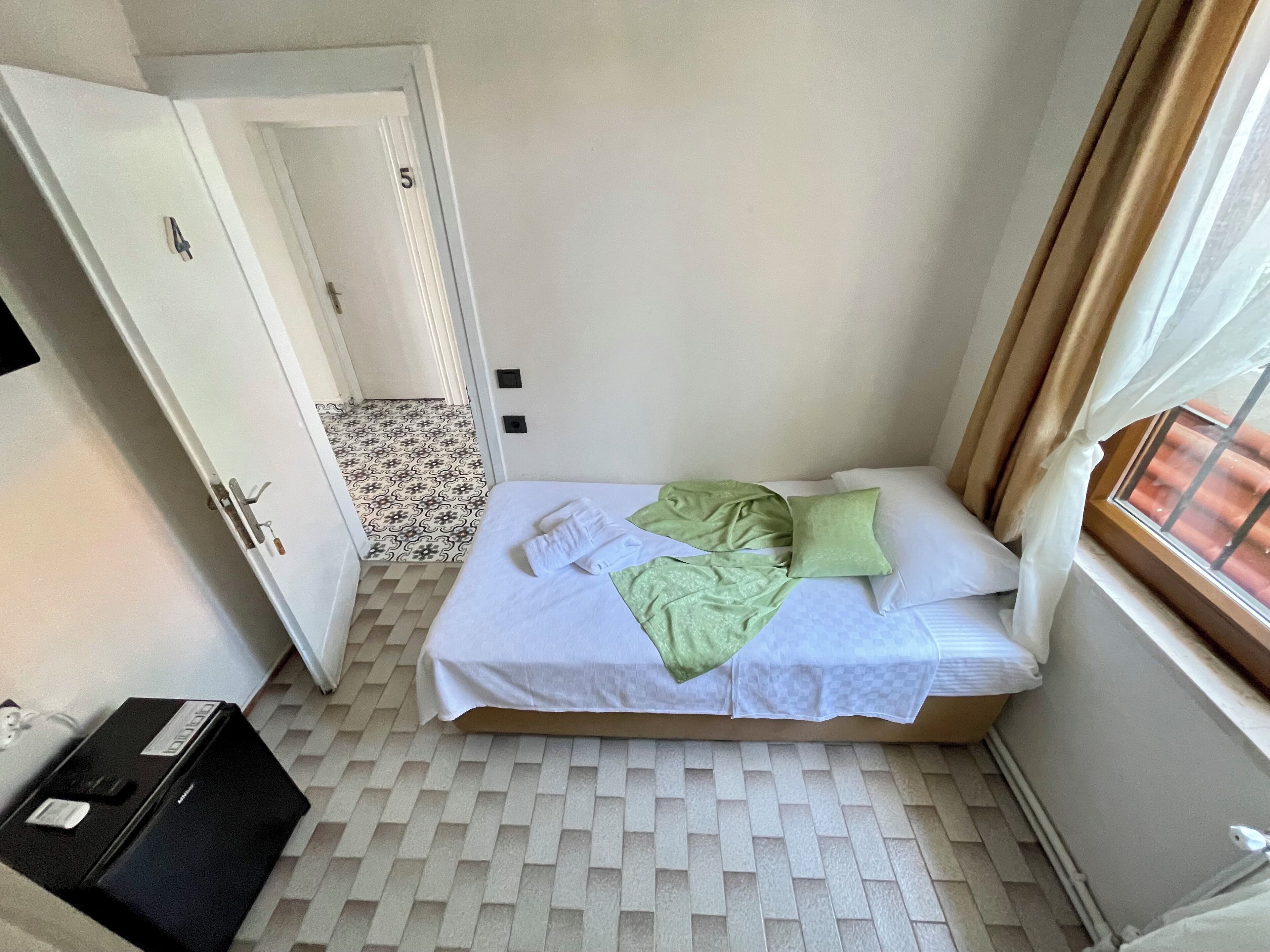A small, neatly arranged bedroom with a bed, a window with curtains, a door, and a patterned floor.