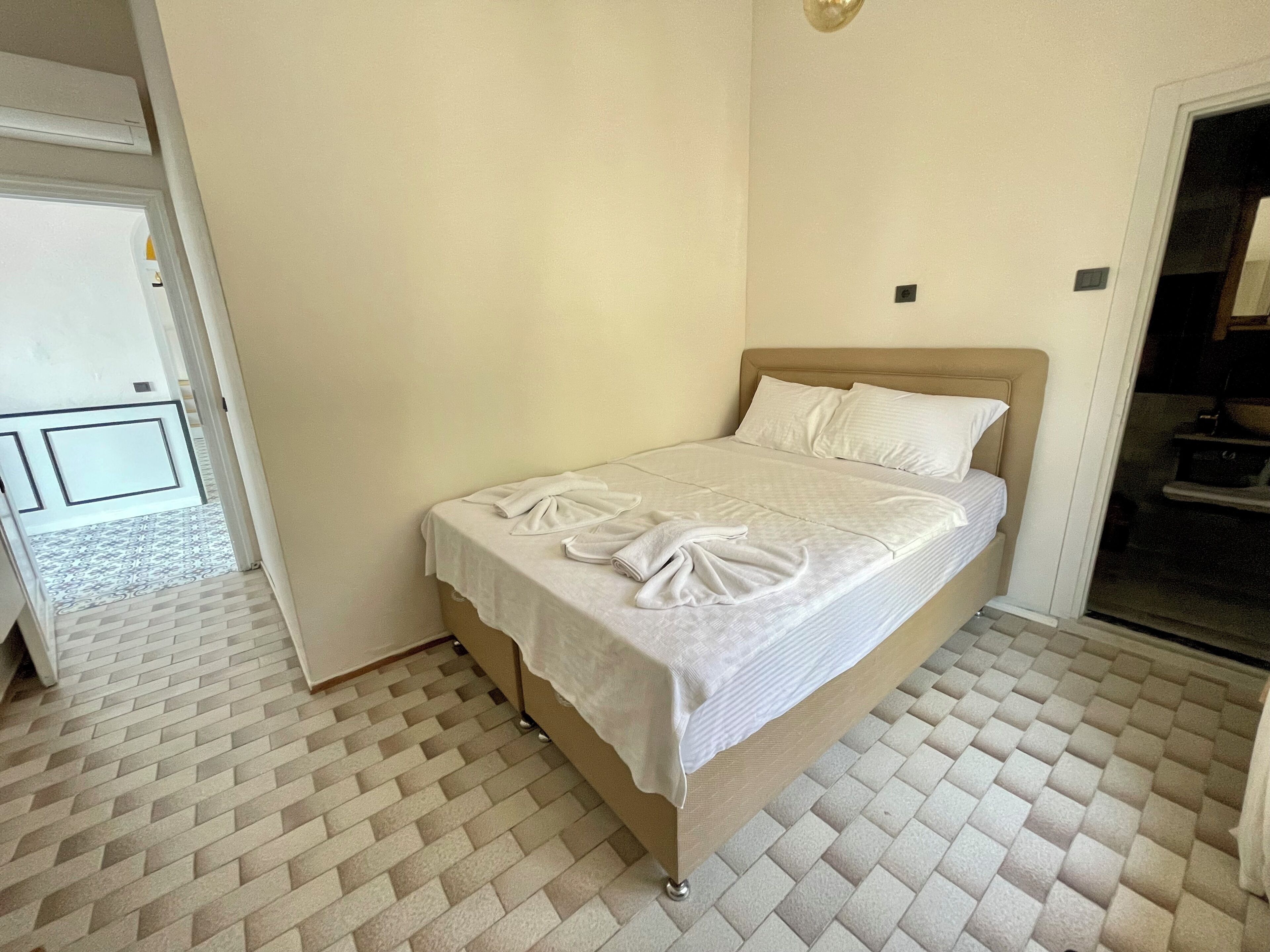 A neatly made bed with a white bedspread and pillows in a room with a herringbone-patterned floor and plain walls.
