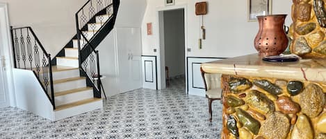Una escalera con barandillas negras y peldaños de madera, un suelo estampado, una pared de piedra con una olla de cobre, y un cuadro enmarcado en la pared.