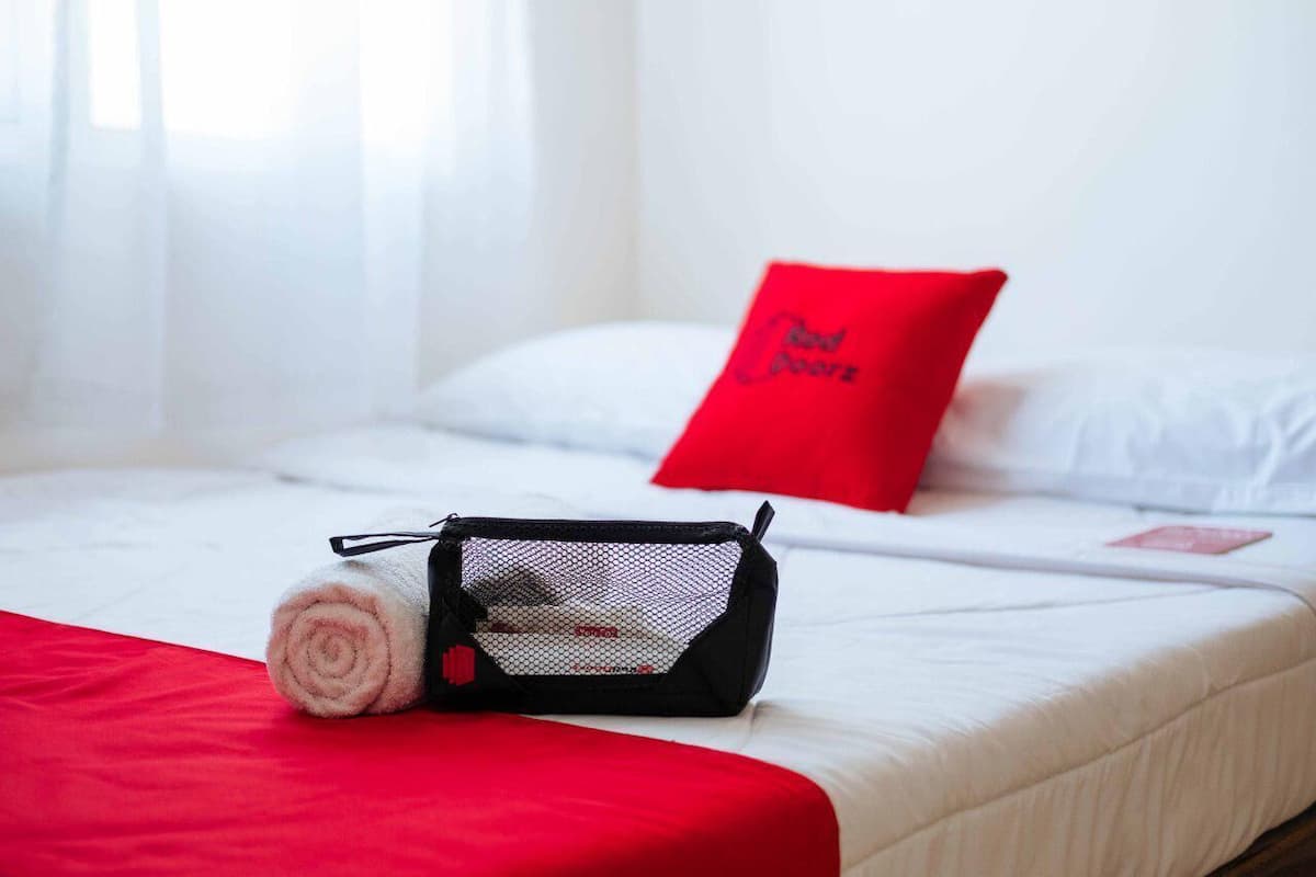 A neatly made bed with a red pillow, a rolled towel, and a black mesh bag on the bed.