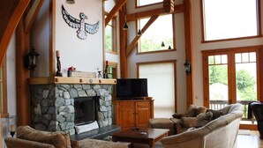 A spacious living room with a stone fireplace, wooden beams, a television, and comfortable seating. - Bay Bluff Haven:  Ocean, Glacier, & Wildlife View (Homer)
