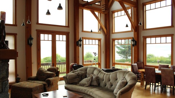 A spacious living room with a large sectional sofa, a dining table with chairs, and multiple windows offering views of the outdoors. - Bay Bluff Haven:  Ocean, Glacier, & Wildlife View (Homer)