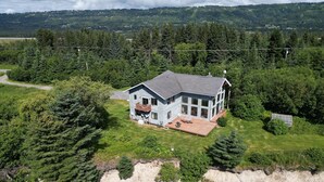 A large house with a deck, surrounded by dense forest and a cliff. - Bay Bluff Haven:  Ocean, Glacier, & Wildlife View (Homer)
