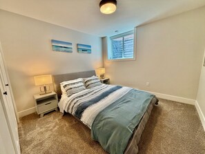 A bedroom with a bed, two bedside tables with lamps, a window with blinds, and two wall-mounted paintings. - New White Rock Suite Close to Pier and Border (Surrey)