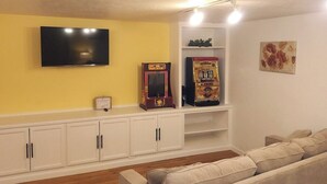 En stue med en beige sofa, et fladskærms-tv, en arkadespilautomat fra en ældre tid og en lille hylde med dekorative genstande.