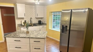 A kitchen with white cabinets, a granite countertop, a stainless steel refrigerator, and a window with a view of greenery. - Bring everyone together while enjoying the privacy of three separate living spaces. This unique Medford retreat features a spacious main home, two private guest homes, a seasonal pool, hot tub, fully equipped kitchens, and room for the whole family. (Medford)