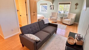 A spacious living room with a dark grey sofa, two armchairs, a small black side table, and a large window with a view of the outdoors. - Bring everyone together while enjoying the privacy of three separate living spaces. This unique Medford retreat features a spacious main home, two private guest homes, a seasonal pool, hot tub, fully equipped kitchens, and room for the whole family. (Medford)