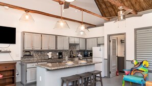 Eine moderne Küche mit Granitarbeitsplatten, weißen Schränken und einer zentralen Kochinsel mit Barhocker. Es gibt Pendelleuchten, einen Deckenventilator und einen an der Wand befestigten Fernseher.