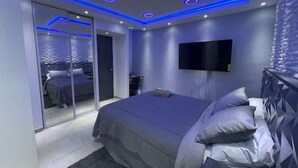 A modern bedroom with a large bed, a TV mounted on the wall, and a sliding glass door leading to a balcony.