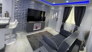 A modern living room with a flat-screen TV, grey sofa, and a white bar stool.
