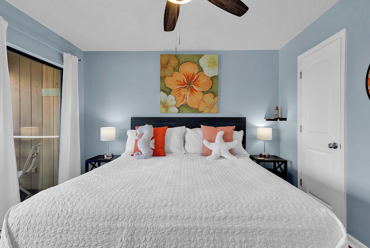 A bedroom with a large bed, two bedside tables with lamps, a ceiling fan, and a door.