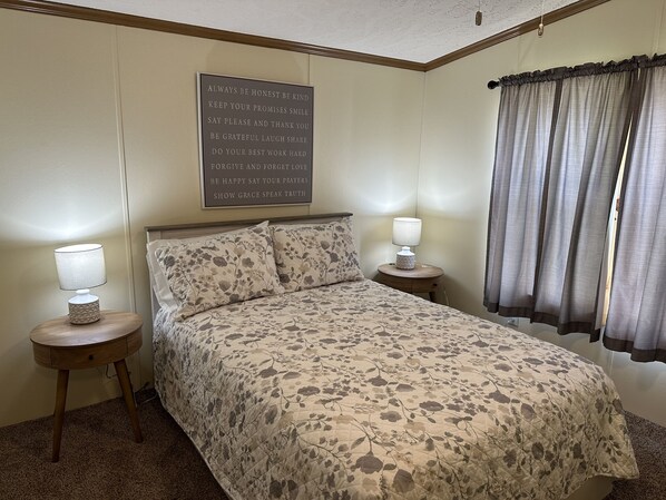 Ein Schlafzimmer mit einer blumigen Tagesdecke, zwei Nachttischlampen, einem an der Wand befestigten Schild mit positiven Aussagen und einem Fenster mit Vorhängen.