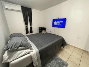 A bedroom with a bed, a TV mounted on the wall, and a window with curtains. - Rincón frente al lago Levittown (Toa Baja Levittown)