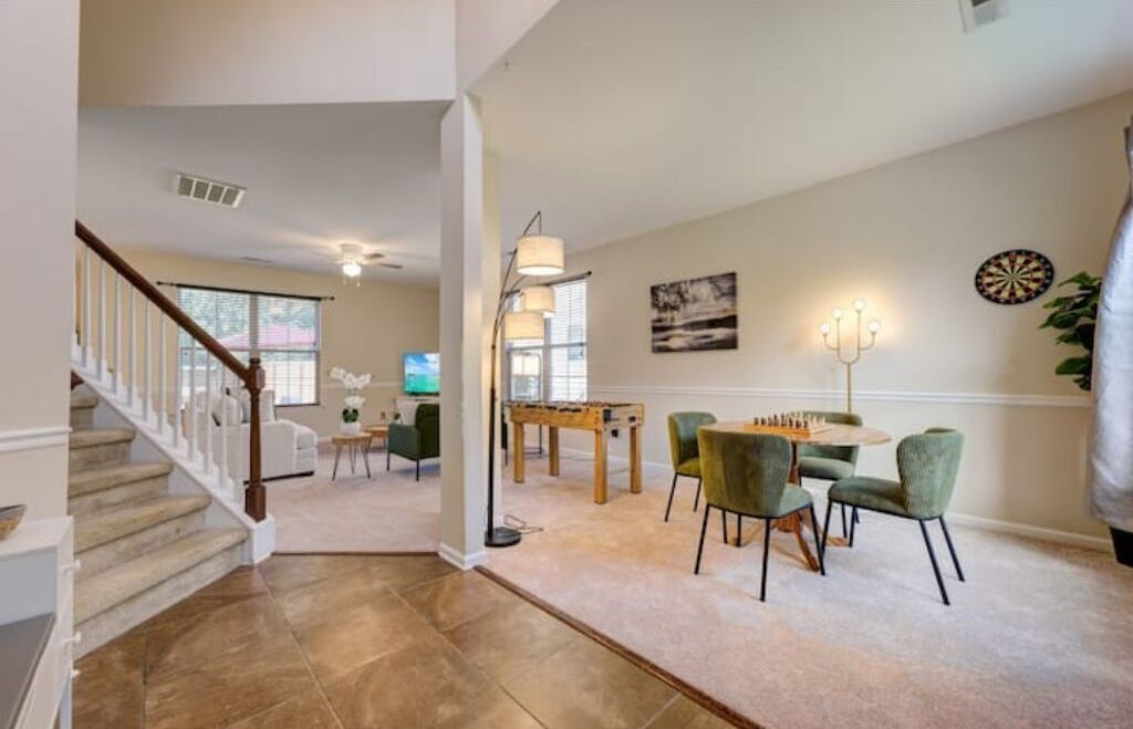 Un salon spacieux avec un escalier, un coin repas avec des chaises vertes et un jeu de fléchettes accroché au mur.