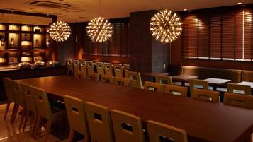 A dining area with a long wooden table and multiple chairs, overhead spherical light fixtures, and a wall with vertical blinds.