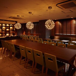A dining area with a long wooden table and multiple chairs, overhead spherical light fixtures, and a wall with vertical blinds. - Super Hotel Mito Natural Onsen (Mito)