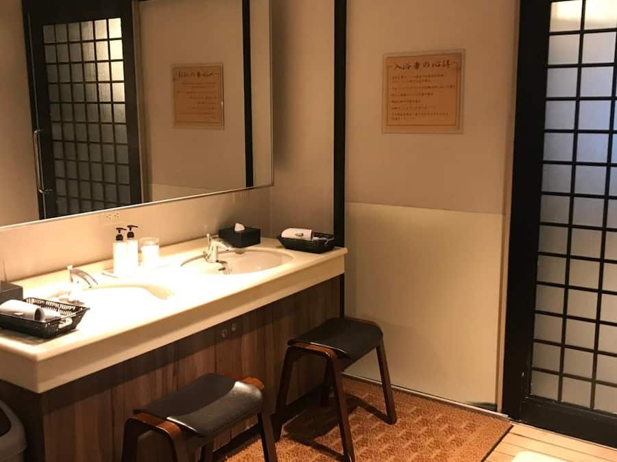 A bathroom with a large mirror, two sinks, a towel rack, and a small stool.