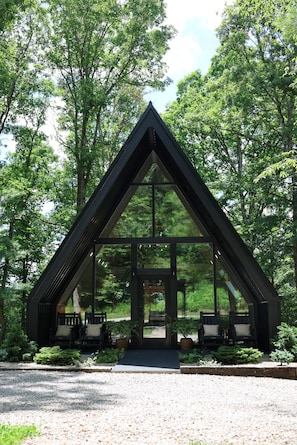 A modern, triangular glass structure with seating outside, surrounded by trees.