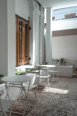 A covered outdoor seating area with white metal chairs and tables, wooden shutters, and a white brick wall.