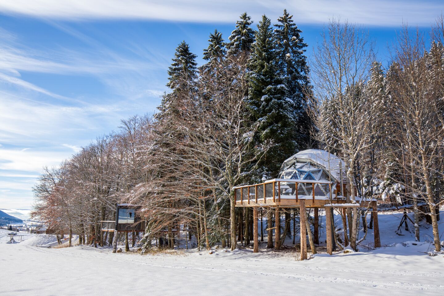 A snow-covered landscape with a glass dome structure, wooden deck, and a small building.