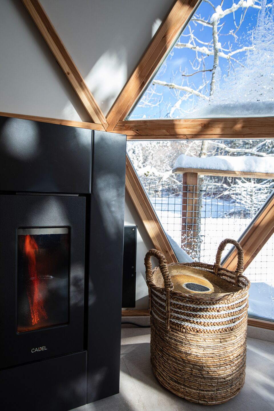A modern interior with a wood-burning stove, a woven basket, and a view of a snowy landscape through a skylight.