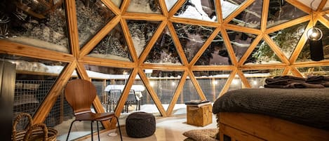 Un interior acogedor de diseño geométrico con una cama, zona de estar y vistas a un paisaje nevado a través de amplios ventanales.