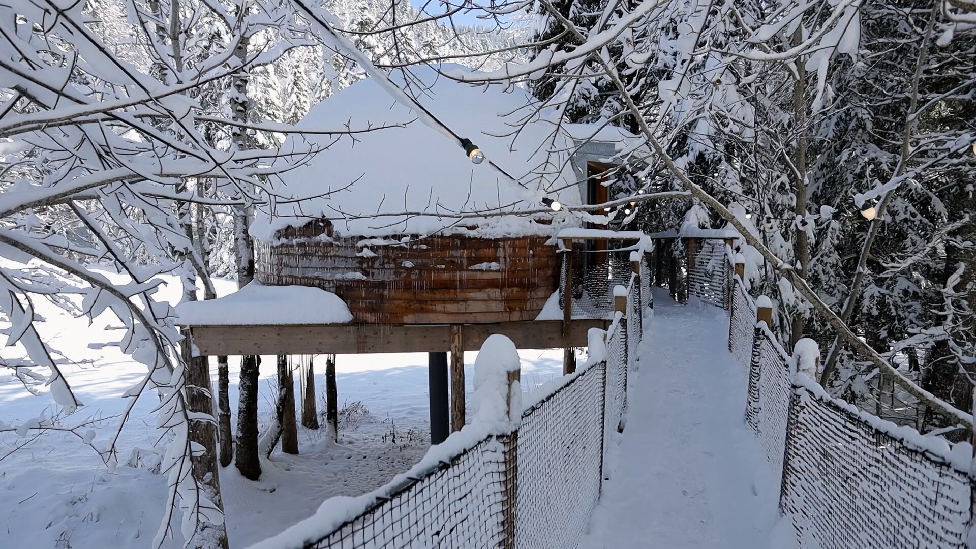 A snow-covered wooden bridge with a metal railing, leading to a building with a snow-covered roof.