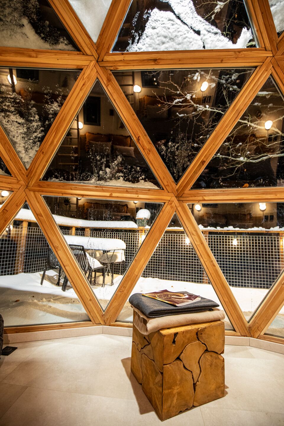Un espacio exterior cubierto de nieve con un banco de madera y un libro, visto a través de una estructura de madera geométrica.