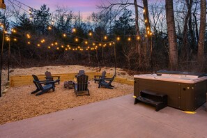 An outdoor seating area with Adirondack chairs, a hot tub, and string lights against a forest backdrop.