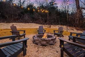 Un foyer entouré de fauteuils Adirondack, avec des guirlandes lumineuses suspendues au-dessus et une clôture en bois en arrière-plan.
