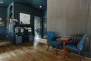 A room with a dark blue wall, a wooden floor, a blue upholstered chair, a small wooden table, and a cabinet with glassware.
