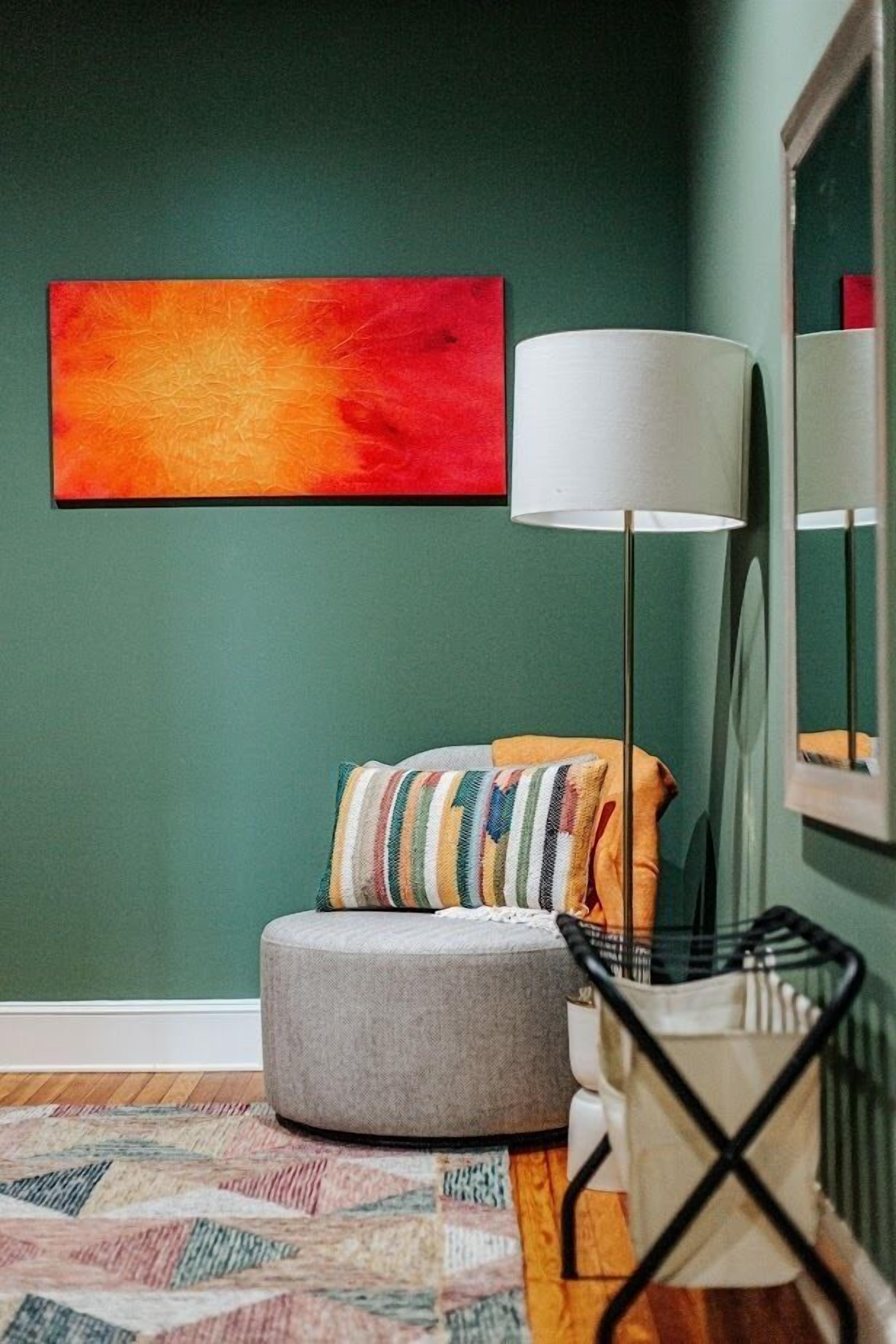 A room with a green wall, a large abstract painting, a grey ottoman, a floor lamp, and a patterned rug.