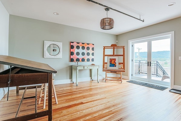 Une pièce spacieuse avec un plancher en bois, une table à manger, un bureau blanc et un chevalet sur lequel est posée une toile. Une grande fenêtre offre une vue sur l’extérieur.