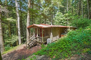 A rustic cabin in a forest with a wooden exterior, a red roof, and a staircase leading to a porch.