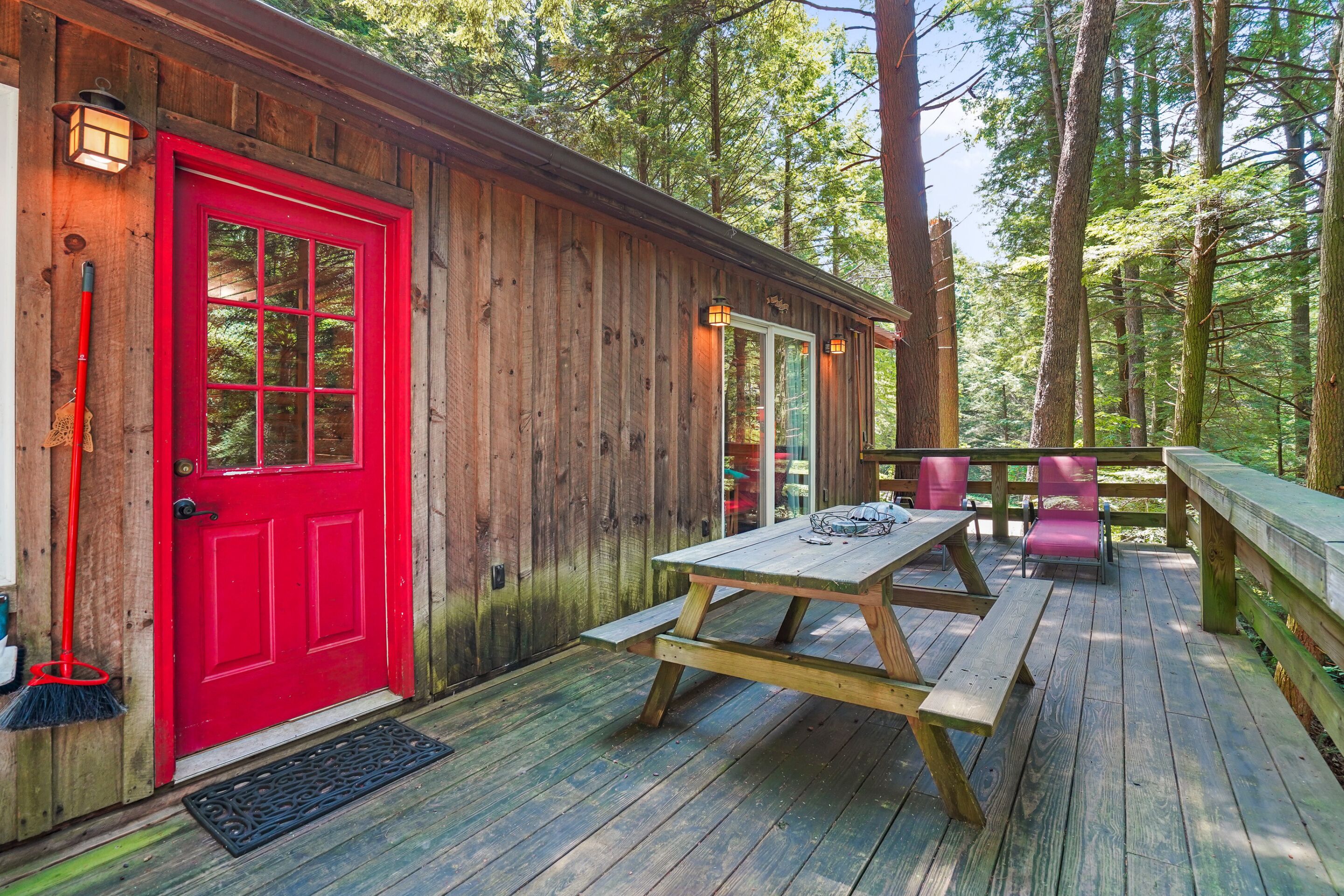 A wooden cabin with a red door, a picnic table, and a deck overlooking a forest.