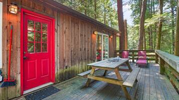 A wooden cabin with a red door, a picnic table, and a deck overlooking a forest.