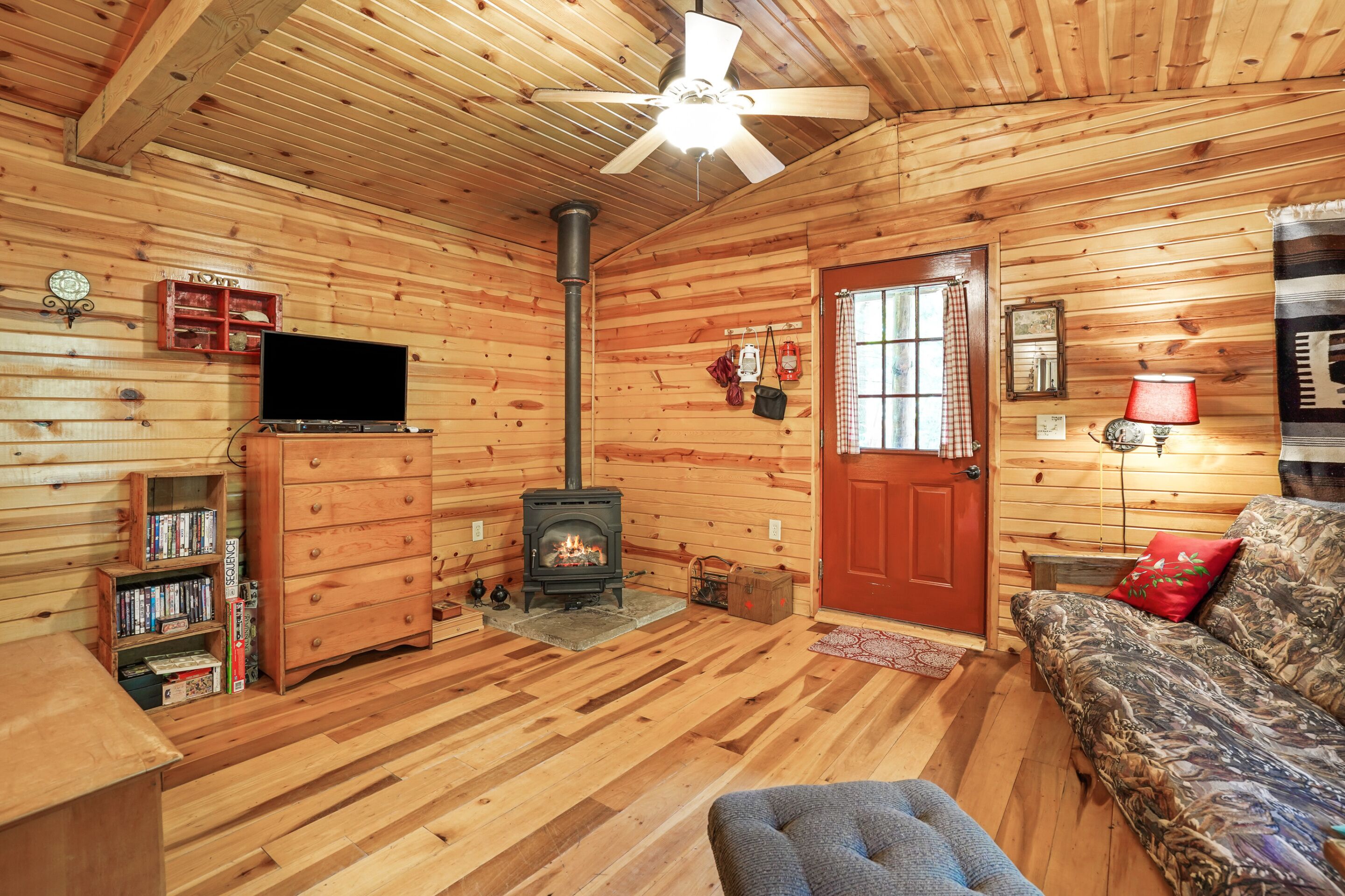 A cozy cabin interior with a fireplace, wooden walls, a ceiling fan, a television, and a couch.