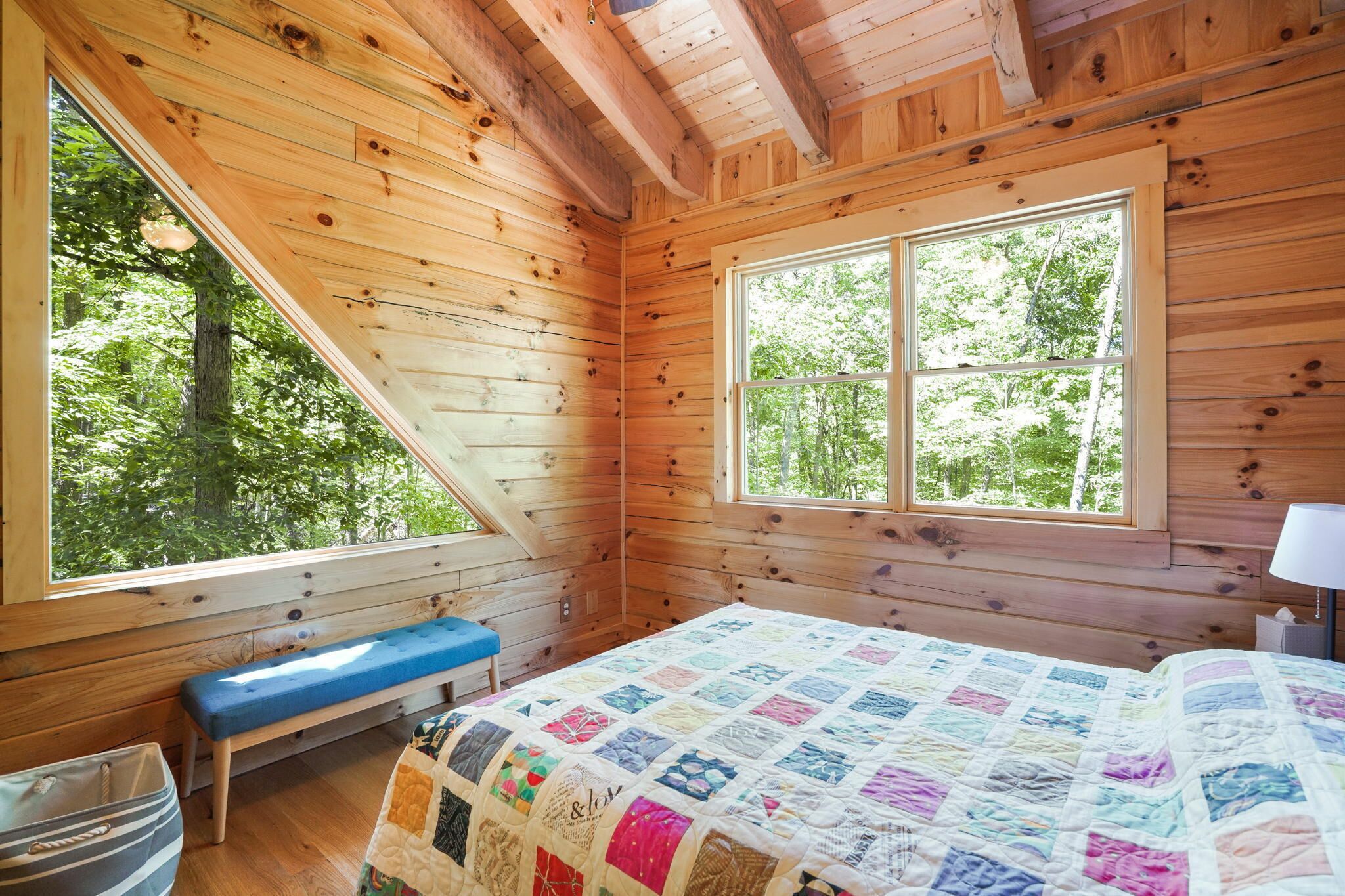 A wooden cabin bedroom with a large window, a bed with a quilt, a bench, and a bedside table.