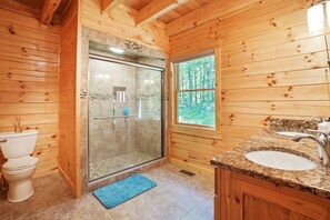 A wooden cabin bathroom with a glass-enclosed shower, a toilet, a sink, and a window with a forest view.