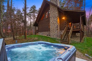 A log cabin with a stone chimney, a hot tub, and a wooden deck.