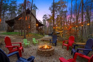 A rustic cabin with a stone exterior, surrounded by a forest. There is a fire pit in the center with several Adirondack chairs around it.