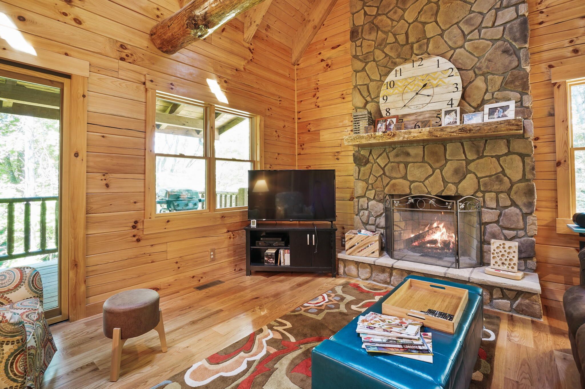 Interior de una cabaña de troncos con chimenea de piedra, paredes de madera, un televisor y una zona de estar acogedora.