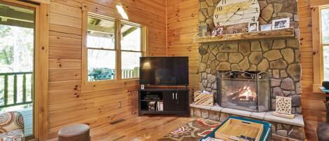 Interior de una cabaña de troncos con chimenea de piedra, paredes de madera, un televisor y una zona de estar acogedora.