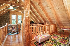 Ein gemütliches Dachzimmer mit Holzbalkendecke, Steinwand, Bücherregal, einer Bank und einem farbenfrohen Teppich.