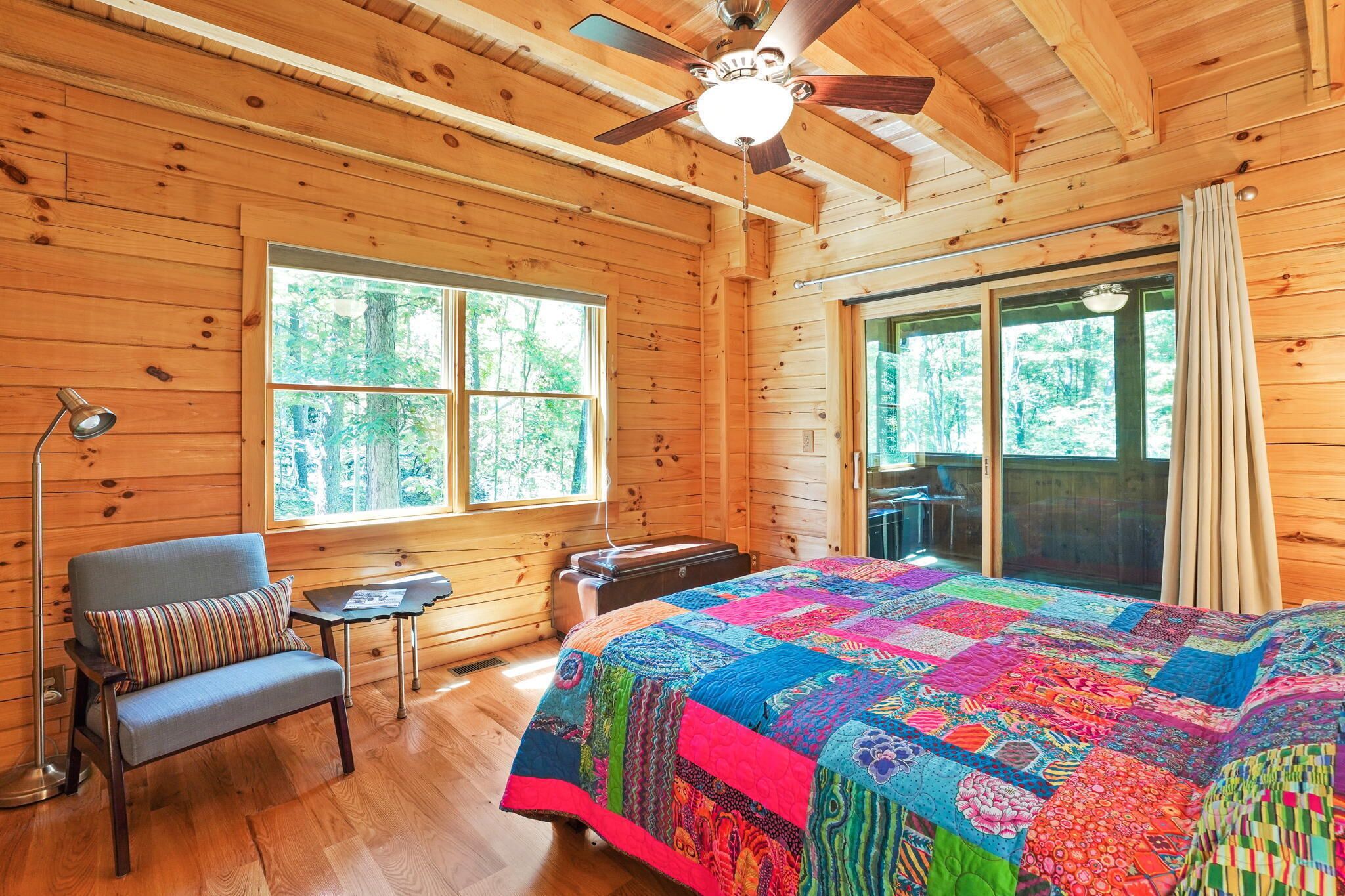 A wooden cabin room with a bed, a chair, a table, and a window.