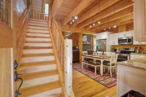 A wooden staircase leading to a kitchen with white cabinets, a dining table, and a colorful rug.