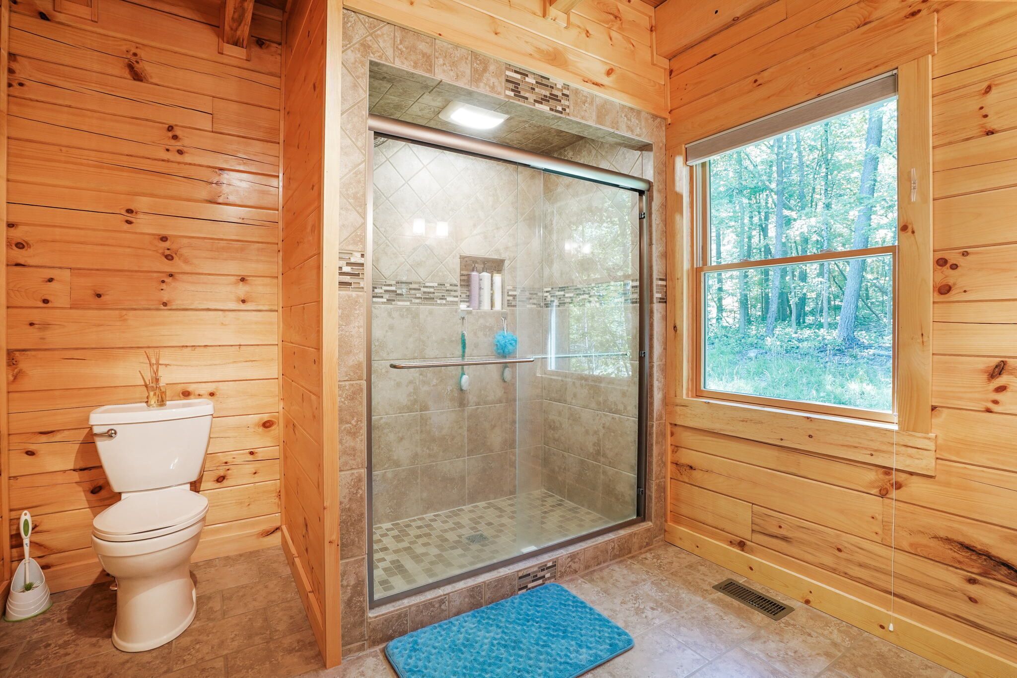 A wooden cabin bathroom with a toilet, a glass-enclosed shower, and a window with a forest view.