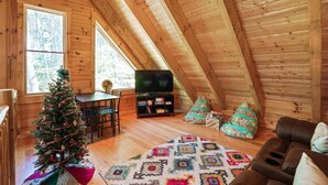 Een gezellig zolderkamertje met een houten plafond, een televisie, een kerstboom en een kamerkleed met dessin.