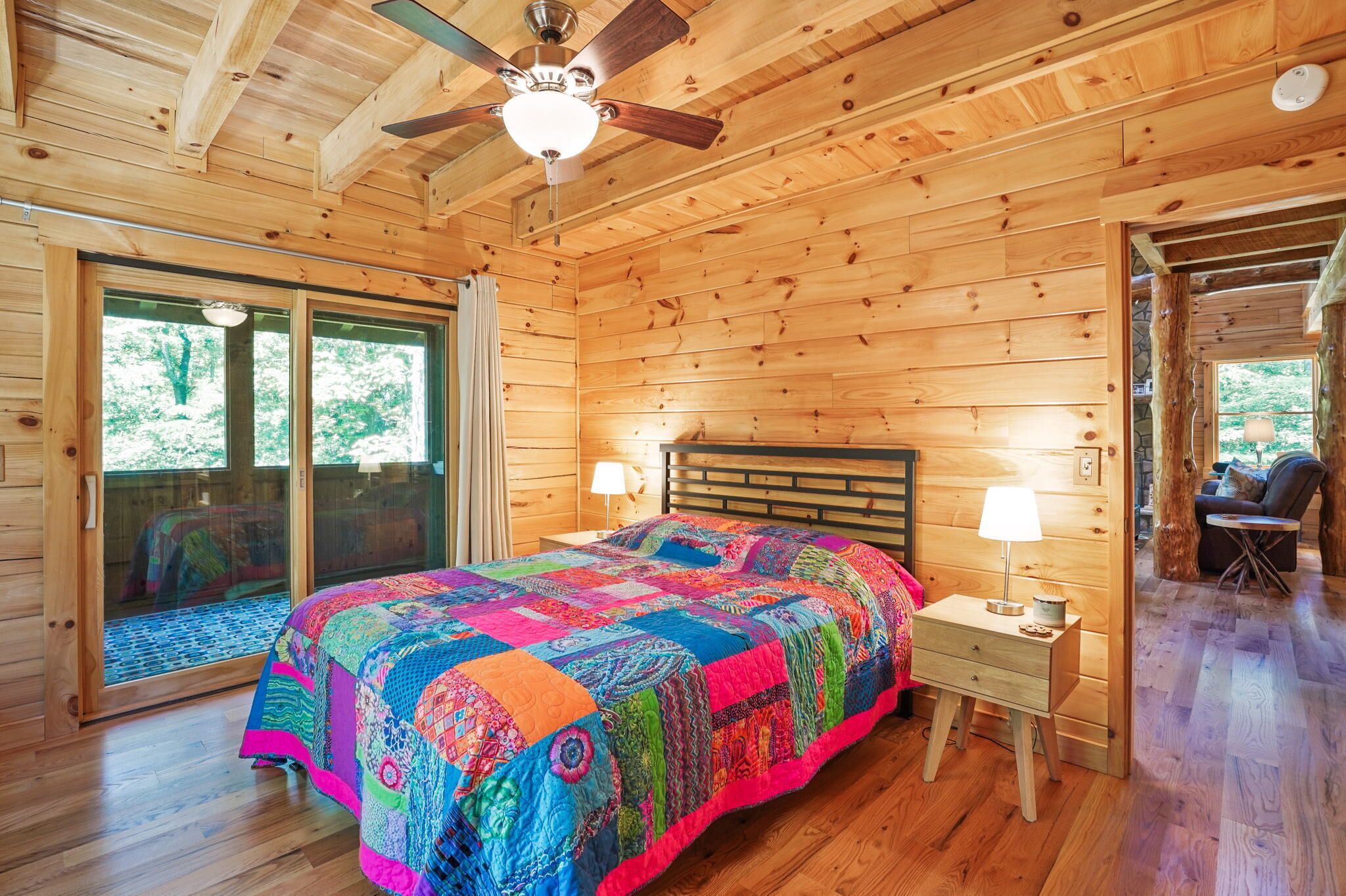 A wooden cabin bedroom with a bed, nightstands, a ceiling fan, and a large window.